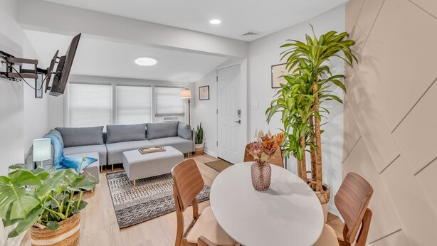 A modern living room with a grey sofa, a dining table with chairs, a flat-screen TV mounted on the wall, and a potted plant.