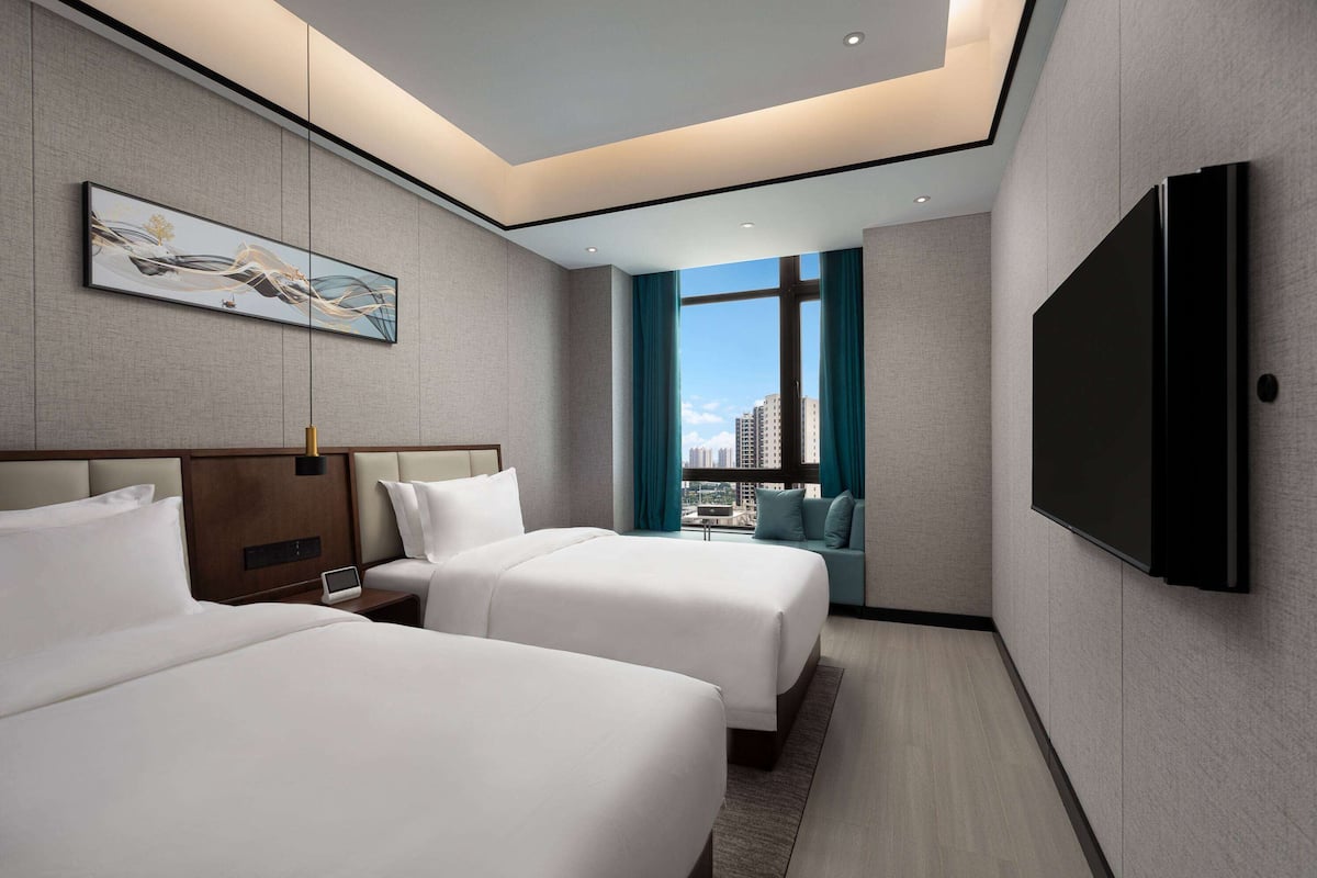 Quarto de hotel com duas camas, televisão de tela plana, um quadro na parede e vista da cidade através de grandes janelas.
