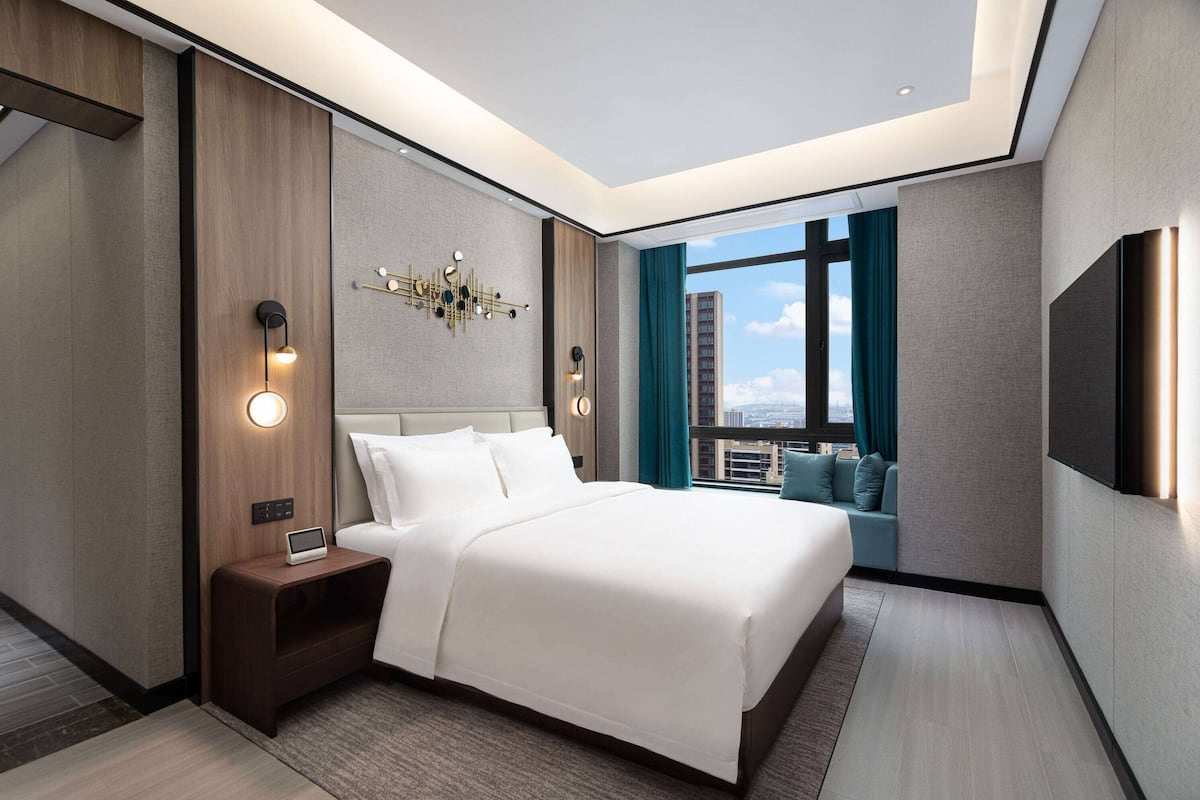 Quarto de hotel moderno com uma cama grande, uma mesa de cabeceira com abajur, TV na parede e vista da cidade através de amplas janelas.