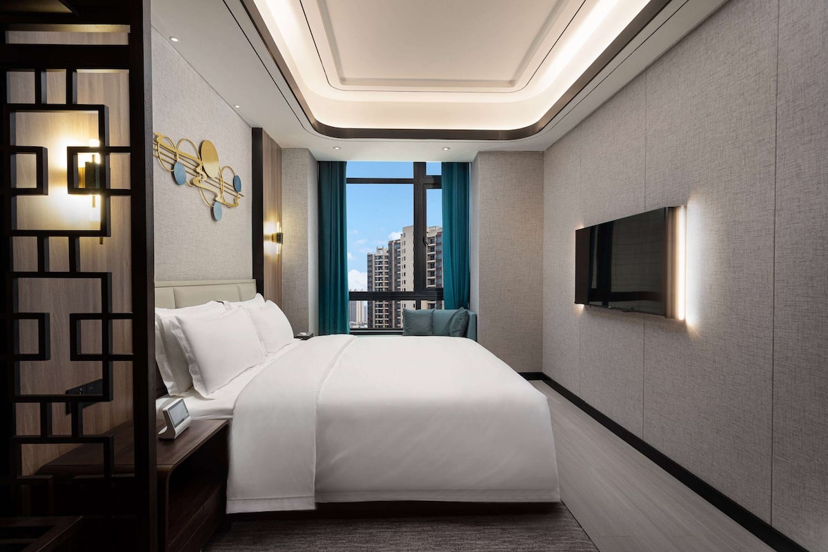 Quarto de hotel moderno com uma cama grande, uma TV fixada na parede e vista para a paisagem urbana pela janela.