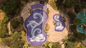 Vista aérea de un parque con una gran fuente serpenteante y un estanque circular más pequeño, rodeados de árboles y senderos.
