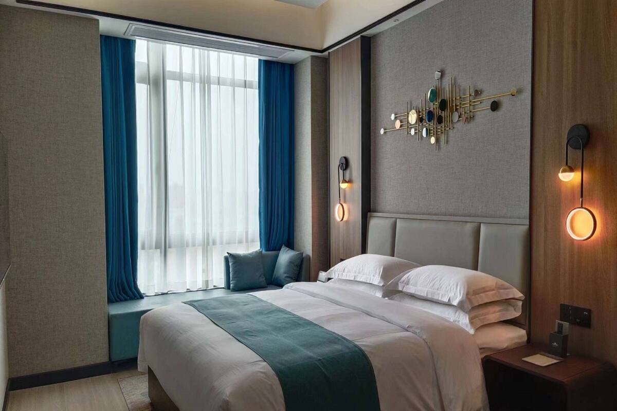 Quarto de hotel moderno com uma cama grande, um espaço de estar junto à janela e um detalhe decorativo na parede.