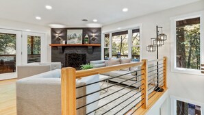 A modern living room with a fireplace, a wooden staircase, and large windows offering a view of the outdoors. - 🌻Have FUN in the Fall Foliage TREETOPS🍁Luxury 4 Suite Cabin🍁Screened Porches🌻 (Berkeley Springs)