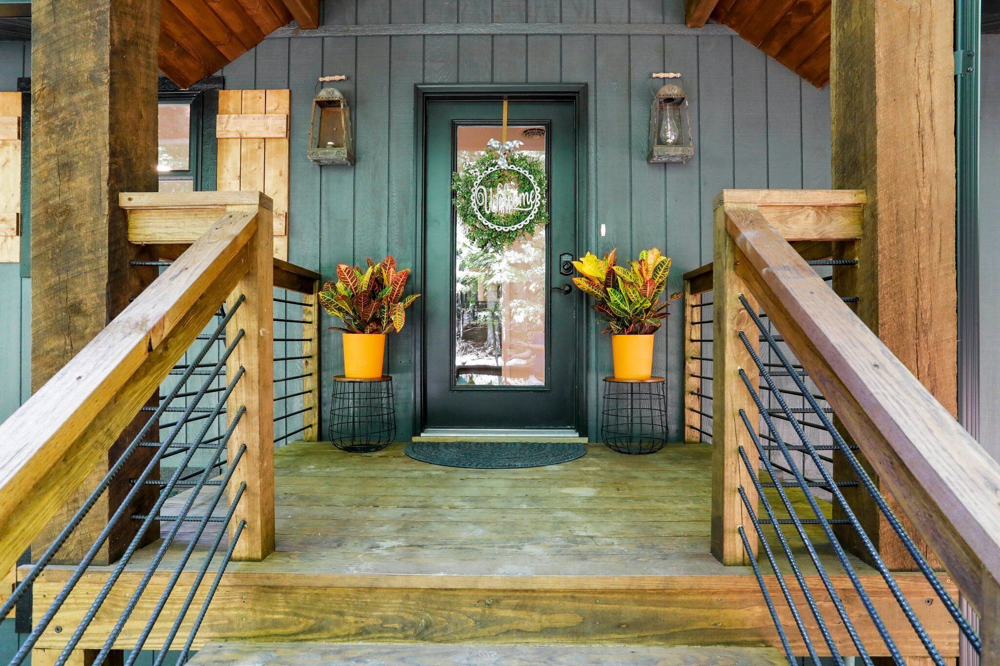 A wooden staircase leading to a dark green door with a wreath, two potted plants, and a welcome sign.