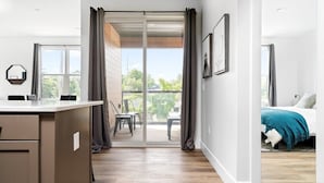 A modern hotel room with a sliding glass door leading to a balcony, a bed with a teal throw, and a desk area with a chair. - 2 King BR I Front Street + Walk DT (Traverse City)