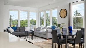Ein modernes Wohnzimmer mit einer Couch, einem Esstisch und Stühlen. Große Fenster sorgen für natürliches Licht und Blick ins Grüne.