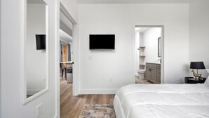 Ein modernes Hotelzimmer mit einem großen Bett, einem flachbildfernseher an der Wand, einem kleinen Nachttisch mit Lampe und Blick in den Küchenbereich.