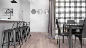 A modern kitchen with a white countertop, black bar stools, and a dining area with a checkered wall and black chairs. - Spacious Condo I Front St + 2 King BR I Near DT (Traverse City)