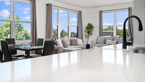 A modern living room with a large window, a sofa, a dining table, and a kitchen area. - Spacious Condo I Front St + 2 King BR I Near DT (Traverse City)