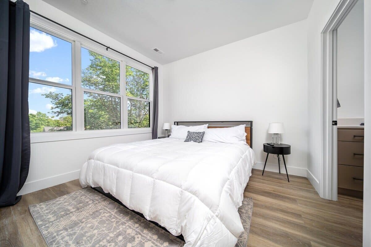 A bedroom with a large bed, a nightstand, a lamp, and a view of greenery through the window.