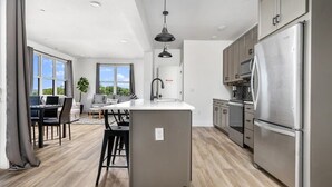 A modern kitchen with a central island, stainless steel appliances, and a dining area with a view. - Spacious Condo I Front St + 2 King BR I Near DT (Traverse City)