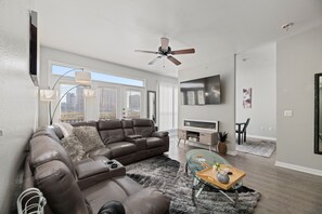 Een moderne woonkamer met een hoekbank, een glazen salontafel, een plafondventilator en een flatscreen televisie.