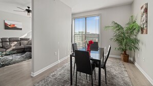 Un salon moderne comprenant un canapé, une table basse en verre, une table à manger avec des chaises, une plante en pot et une grande fenêtre avec des stores.