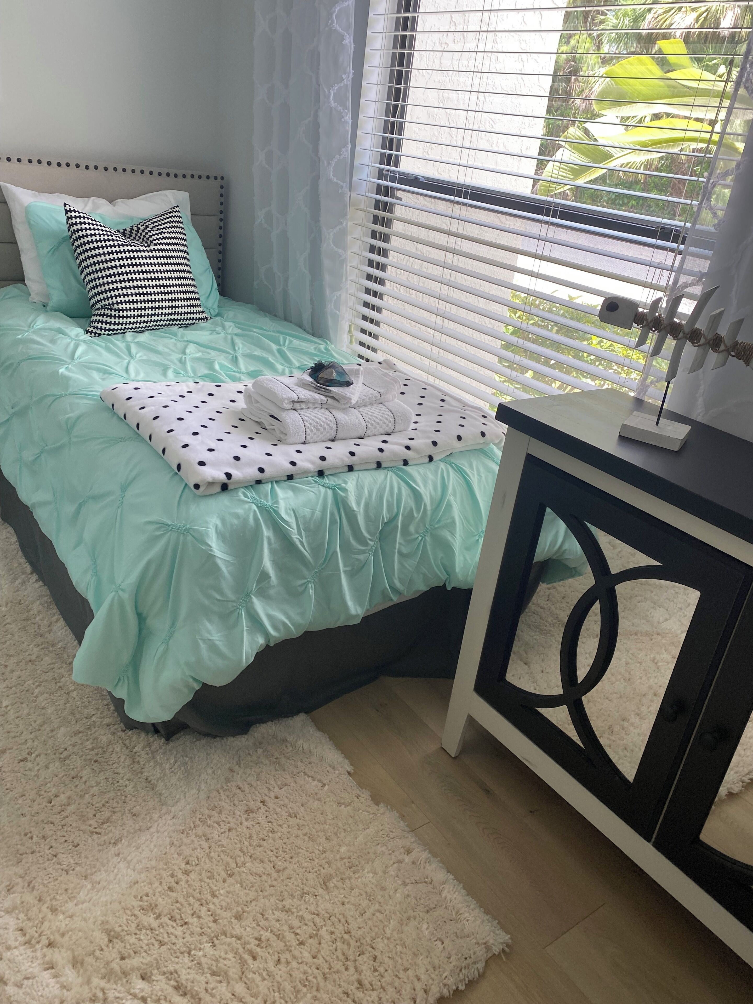 Un lit bien fait, recouvert d’une couvre-lit turquoise et agrémenté d’un coussin à pois, une petite table de chevet ornée d’un objet décoratif, et une vue sur la verdure par la fenêtre.