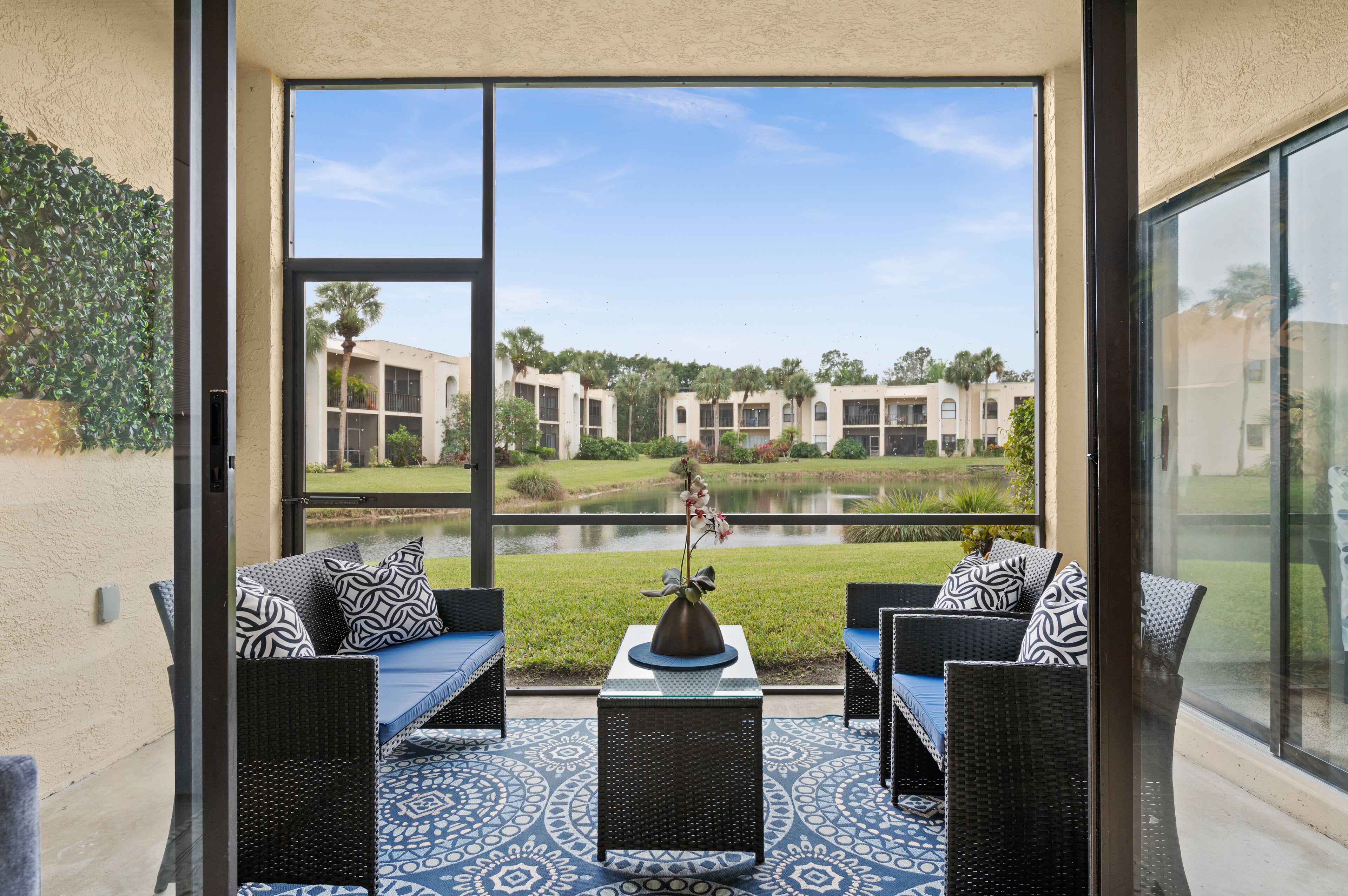 A patio with wicker furniture and a vase, overlooking a building complex and a pond.