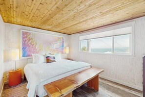 A bedroom with a wooden ceiling, a large bed, a bench, a window with a view, and a colorful abstract painting. - A Serene Waterfront Sanctuary with 360° Views (Bainbridge Island)