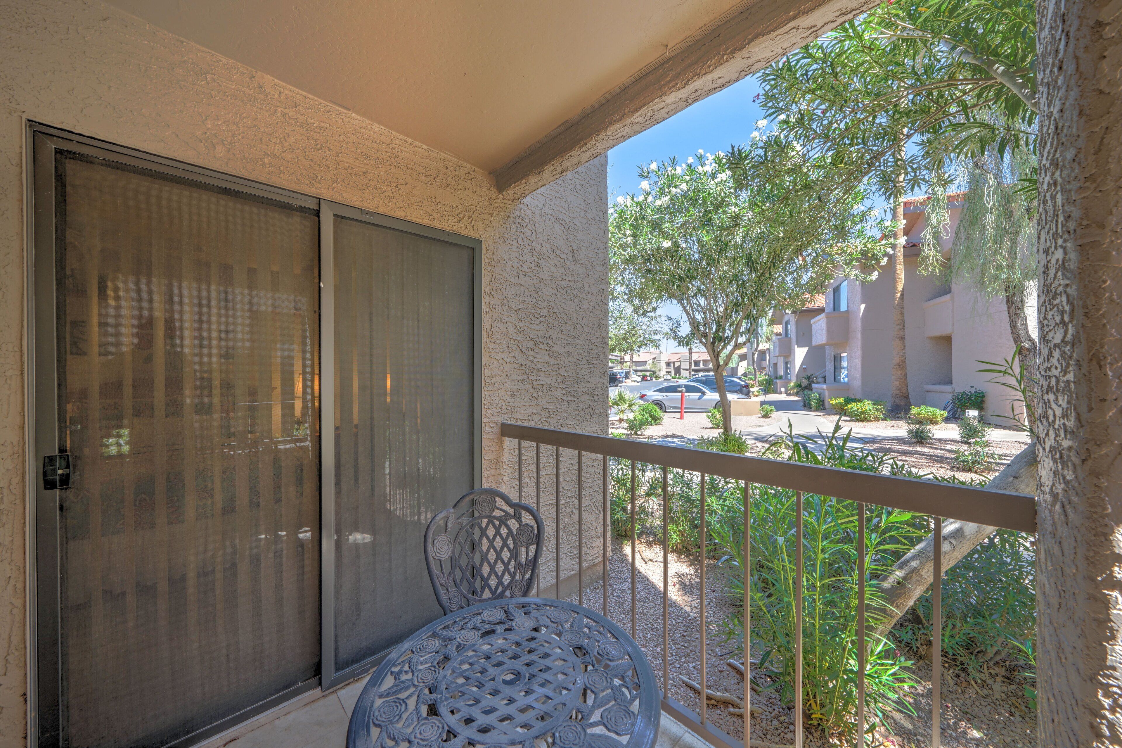 A balcony with a metal chair and a table, overlooking a street with buildings and a tree.