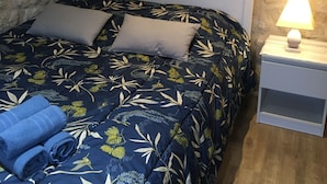A bed with a white headboard, a blue floral comforter, and two pillows. There is a small bedside table with a lamp and a folded blue towel on the bed. - Coquet Studio CDG , Parc Astérix (Coye-la-Forêt)