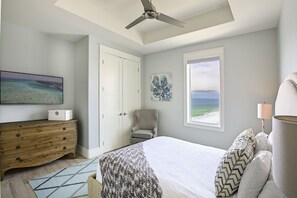 A bedroom with a bed, a dresser, a chair, a ceiling fan, a window with a view of the beach, and artwork on the wall.