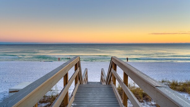Un muelle de madera que lleva a una playa, con una vista despejada del océano y una puesta de sol al fondo.