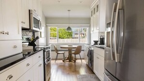 A modern kitchen with white cabinets, stainless steel appliances, a dining area with a round table and chairs, and a view of the outdoors through large windows. - West Boulevard Hideway (Rapid City)