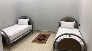 Deux lits simples avec des structures en métal, une literie blanche et un tapis à motifs rouges et beiges sur un sol carrelé.