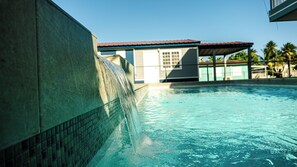 A swimming pool with a water feature, a building with a red roof, and palm trees in the background.