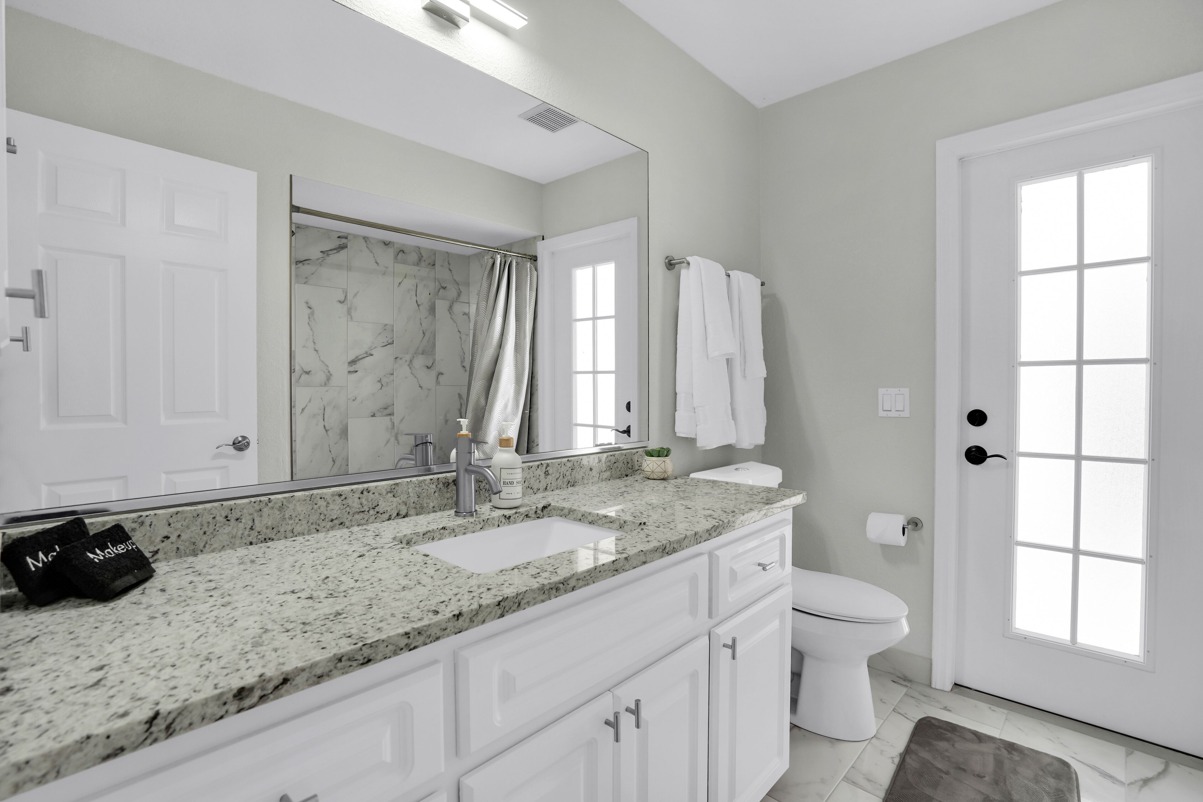 Une salle de bain moderne avec un comptoir en marbre, des meubles blancs, un grand miroir et une douche dotée d’une porte en verre.