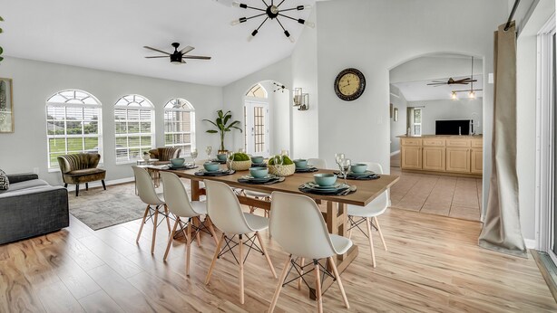 Une salle à manger spacieuse, avec une grande table en bois dressée pour six personnes, entourée de chaises blanches. On y trouve un ventilateur de plafond, une grande horloge au mur et, au fond, une vue sur la cuisine.