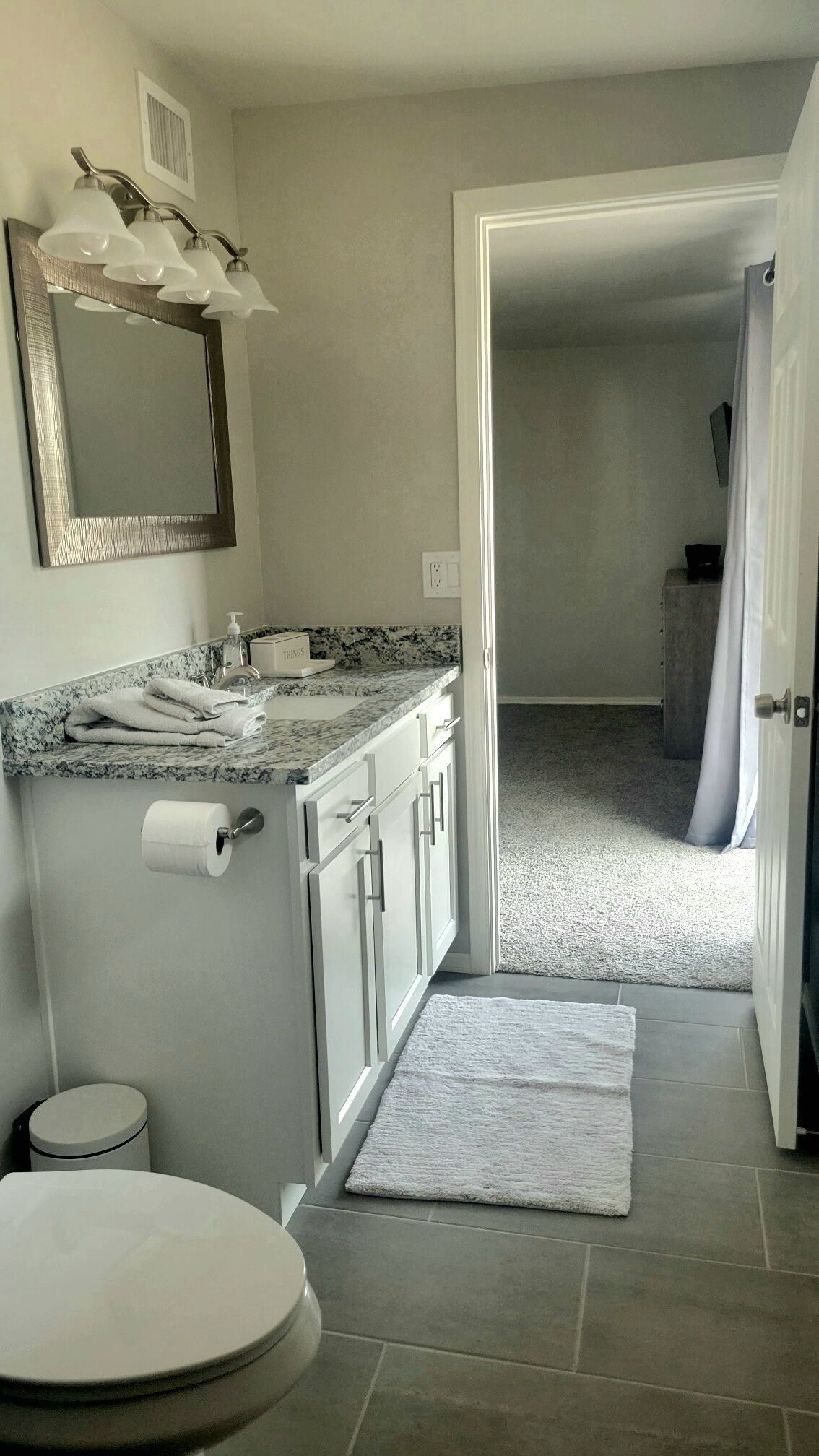 A bathroom with a marble countertop, a toilet, a sink with a mirror, and a shower area visible through an open door.