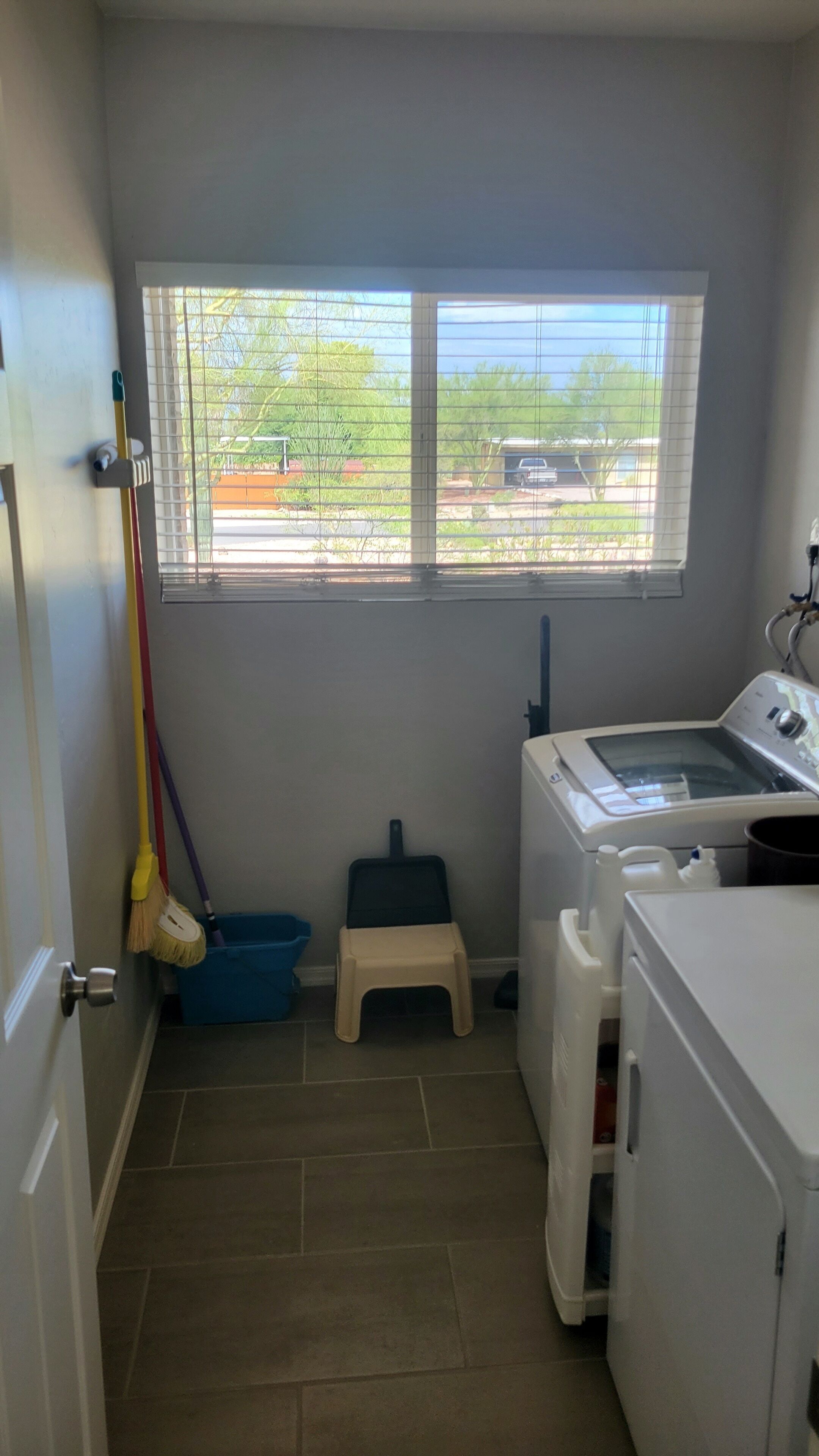 Un cuarto de lavandería con lavadora, un balde para fregar, una escoba y una ventana que da a un estacionamiento y zonas verdes.