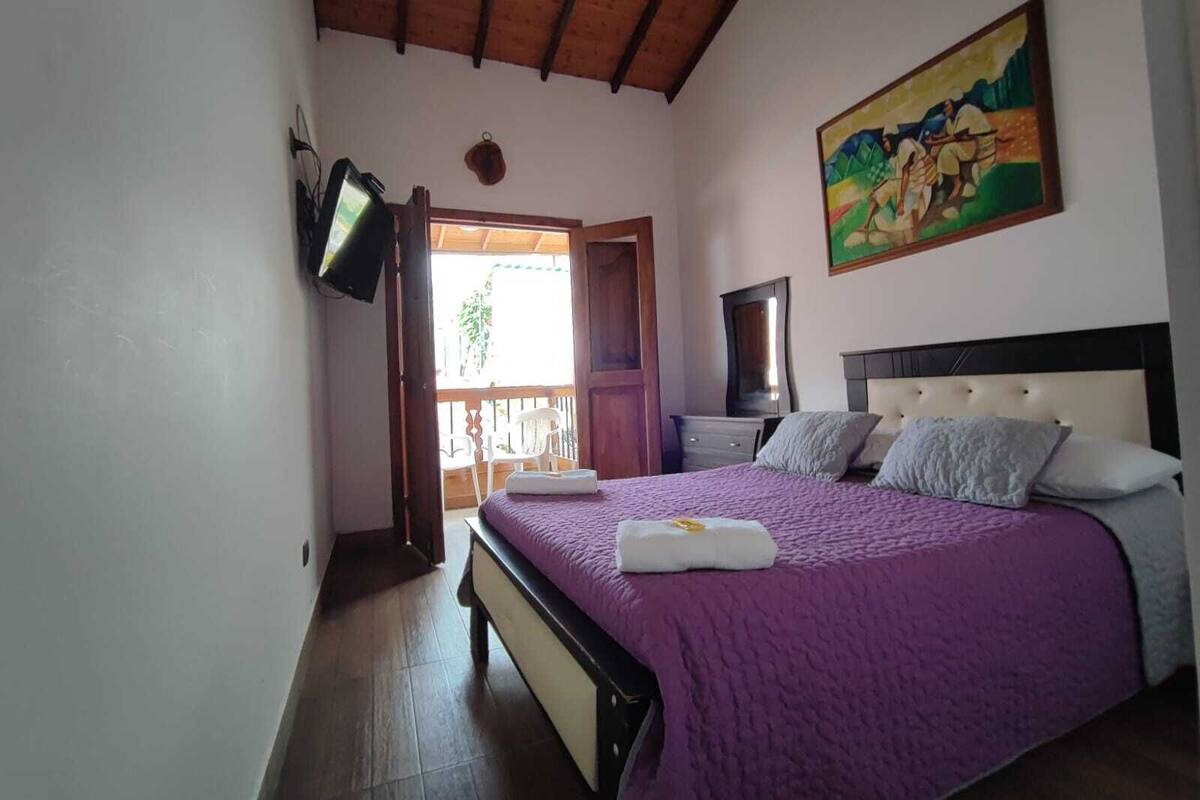 A bedroom with a wooden ceiling, a bed with a purple bedspread, a painting on the wall, and a television on the wall.