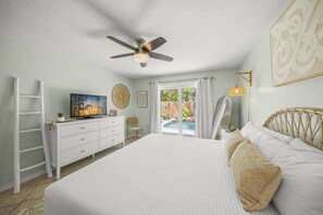 A bedroom with a bed, a ceiling fan, a dresser, a TV, and a view of a pool outside.