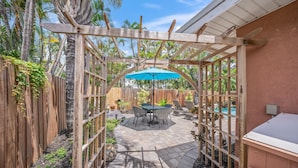 A patio area with a wooden pergola, a table with chairs, and a blue umbrella. - On Turtle Time - Beach Cottage w/ Heated Pool & Close to the Sand (Holmes Beach)