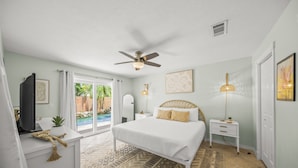 A bedroom with a bed, a ceiling fan, a nightstand, a lamp, and a view of the outdoors. - On Turtle Time - Beach Cottage w/ Heated Pool & Close to the Sand (Holmes Beach)