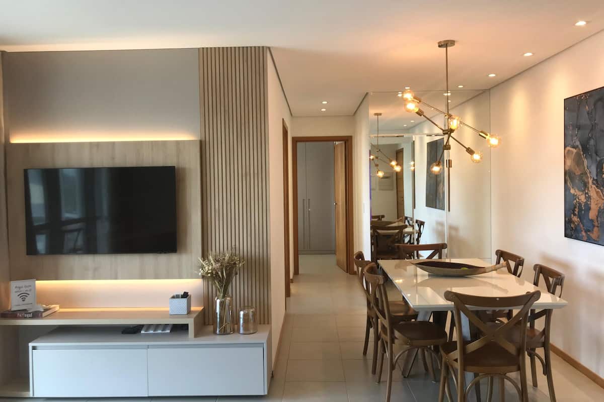Uma sala de estar moderna com televisão de tela plana, um espaço de refeições com cadeiras de madeira e um espelho grande refletindo o ambiente.