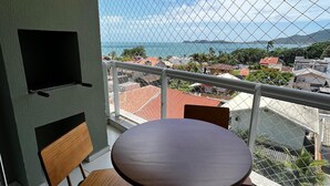 Een balkon met een ronde tafel en twee stoelen, een aan de muur bevestigde televisie, en uitzicht op zee en gebouwen.