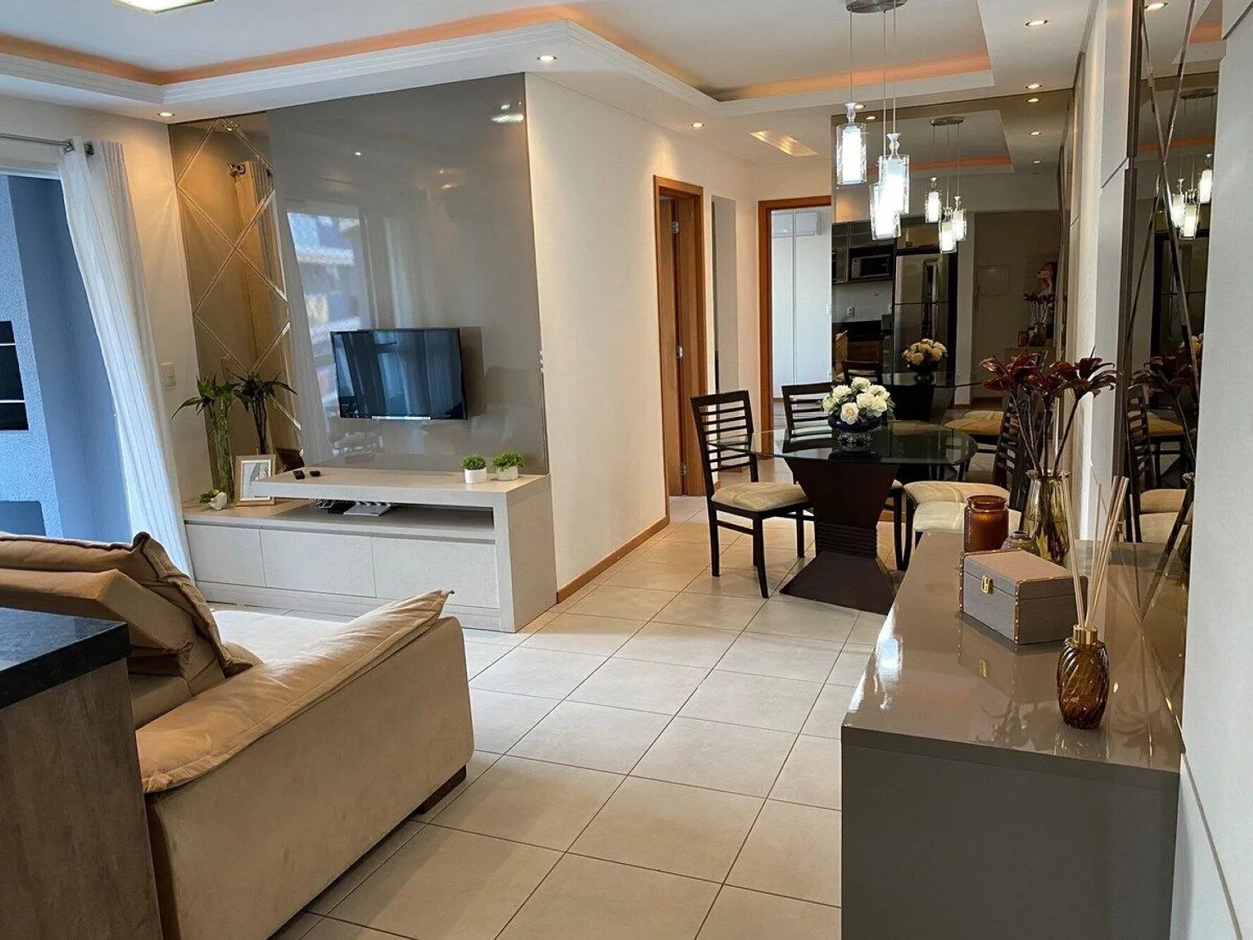 A modern living room with a sofa, a dining table with chairs, a TV mounted on a wall, and a kitchen area visible through an open door.