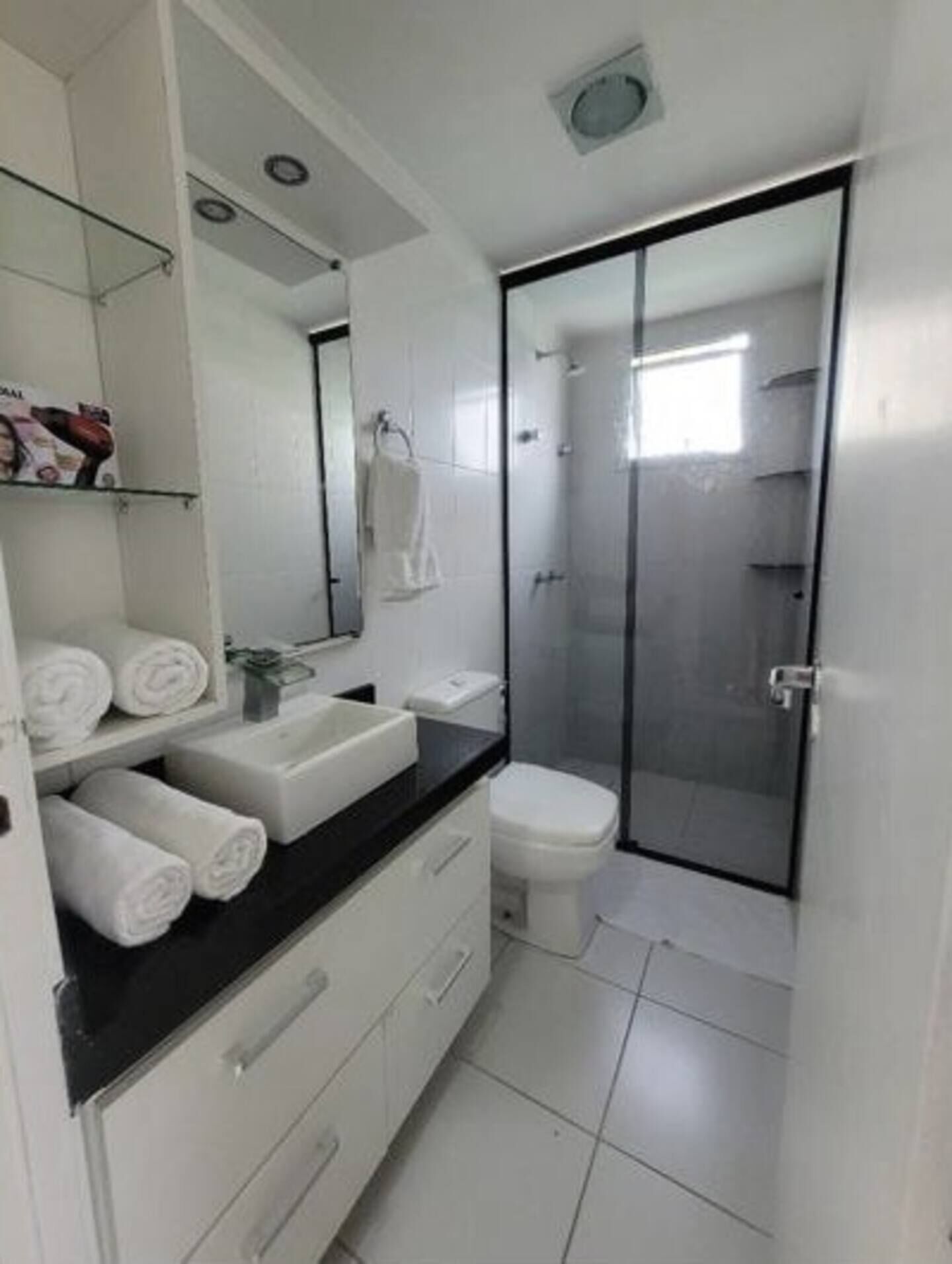 Un baño moderno con lavamanos, inodoro y zona de ducha blancos. Hay toallas enrolladas ordenadamente en un estante.