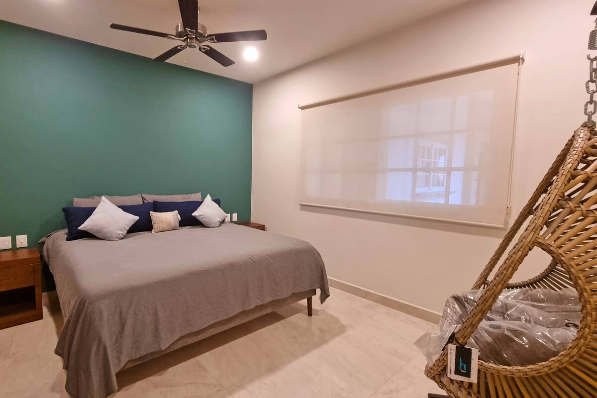 Ein Schlafzimmer mit einem Bett, Nachttischen, einem Deckenventilator, einem Fenster mit Jalousien und einem Korbstuhl.