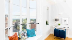Ein heller Raum mit einem großen Fenster, einem blauen Stuhl und einem weißen Schreibtisch mit einer Lampe.