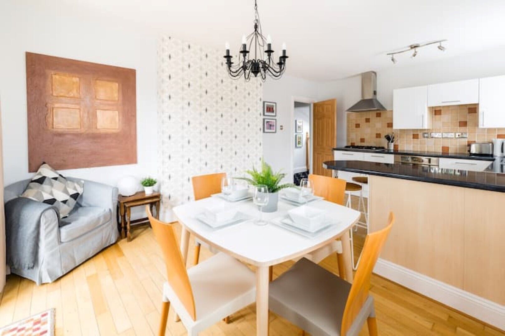 A modern kitchen with a central island, a dining area with a white table and chairs, and a living room with a sofa and a patterned wall.