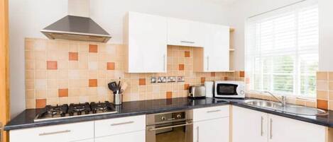 Une cuisine moderne avec des meubles blancs, une cuisinière à gaz, un four à micro-ondes et une fenêtre équipée de stores.