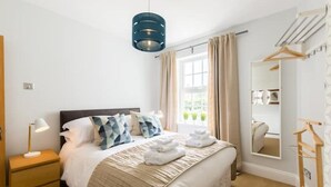 A neatly arranged bedroom with a bed, bedside table, lamp, and a hanging light fixture. - Malvern Wells Apartment with Lovely Balcony Views (Malvern)