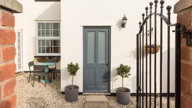 A white building with a grey door, a small table with chairs, and two potted plants. - Fabulous 3 Bed Apartment with Private Courtyard (Malvern)