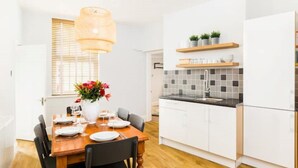 A modern kitchen with white cabinets, a wooden dining table, and a hanging light fixture. - Fabulous 3 Bed Apartment with Private Courtyard (Malvern)