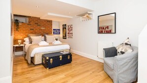 A bedroom with a brick wall, a bed, a dresser, a suitcase, and a gray sofa. - Fabulous 3 Bed Apartment with Private Courtyard (Malvern)
