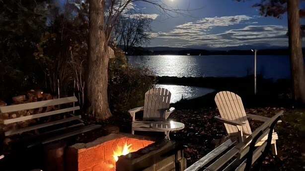 Un espacio al aire libre con sillas Adirondack y un brasero, con vista a un lago bajo la luz de una luna llena.