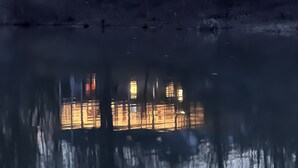 Una casa con un amplio ventanal que se refleja en el agua, rodeada de árboles.