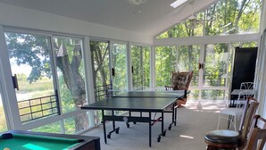 Una mesa de billar con superficie de fieltro verde, una vara de billar y una bola blanca. Hay cuatro sillas de madera con asientos acolchados. La habitación tiene piso alfombrado y ventanas grandes con vistas a árboles y un edificio.